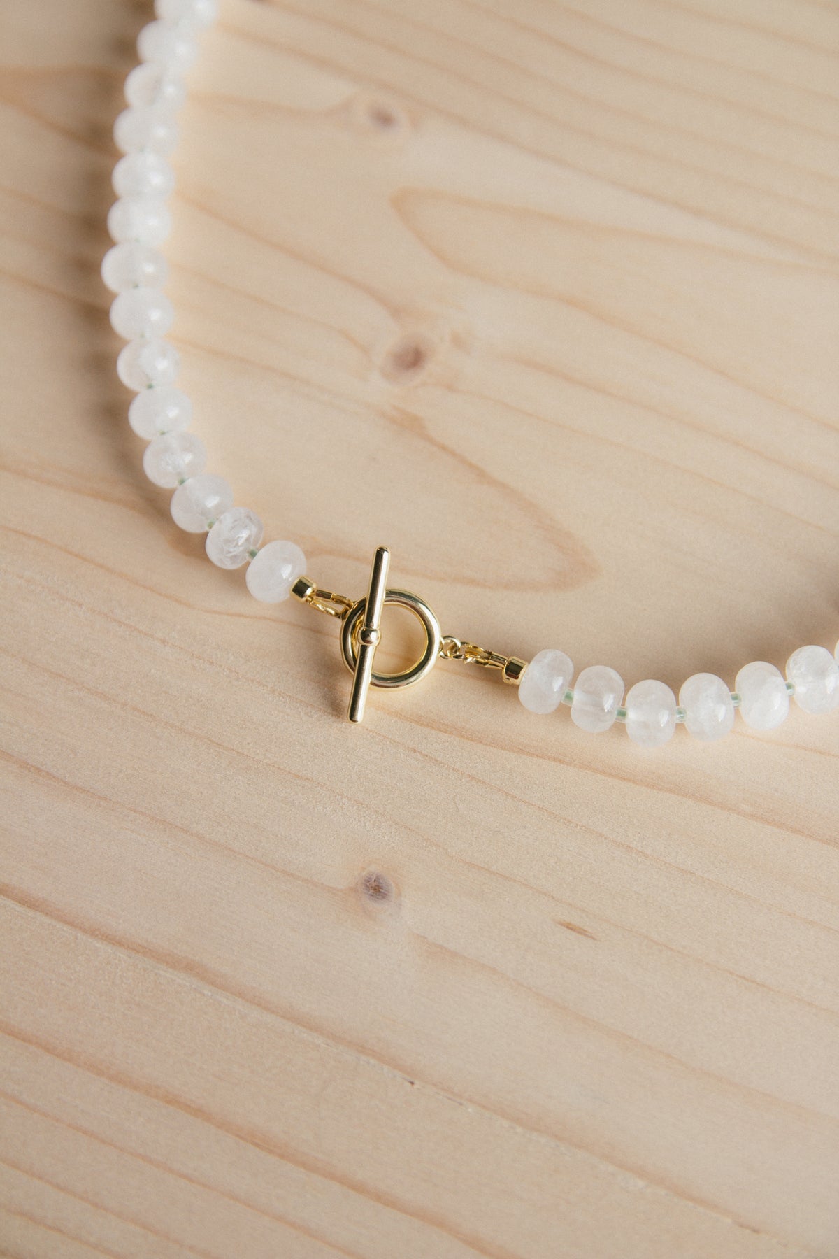 A close-up of a beaded necklace with small white faceted beads and a gold toggle clasp, laid on a light wood surface.