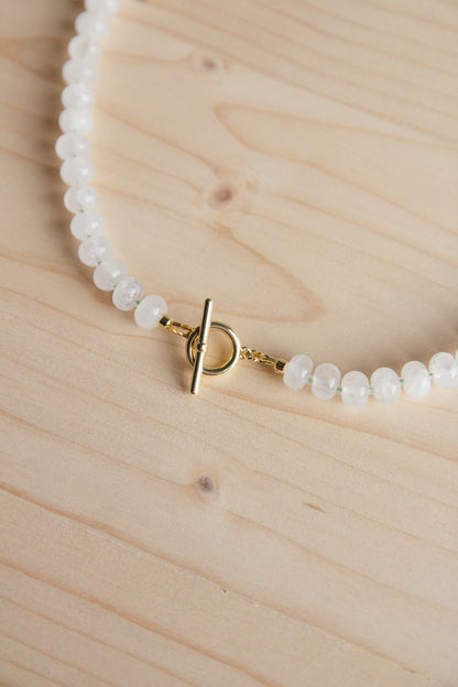A close-up of a beaded necklace with small white faceted beads and a gold toggle clasp, laid on a light wood surface.