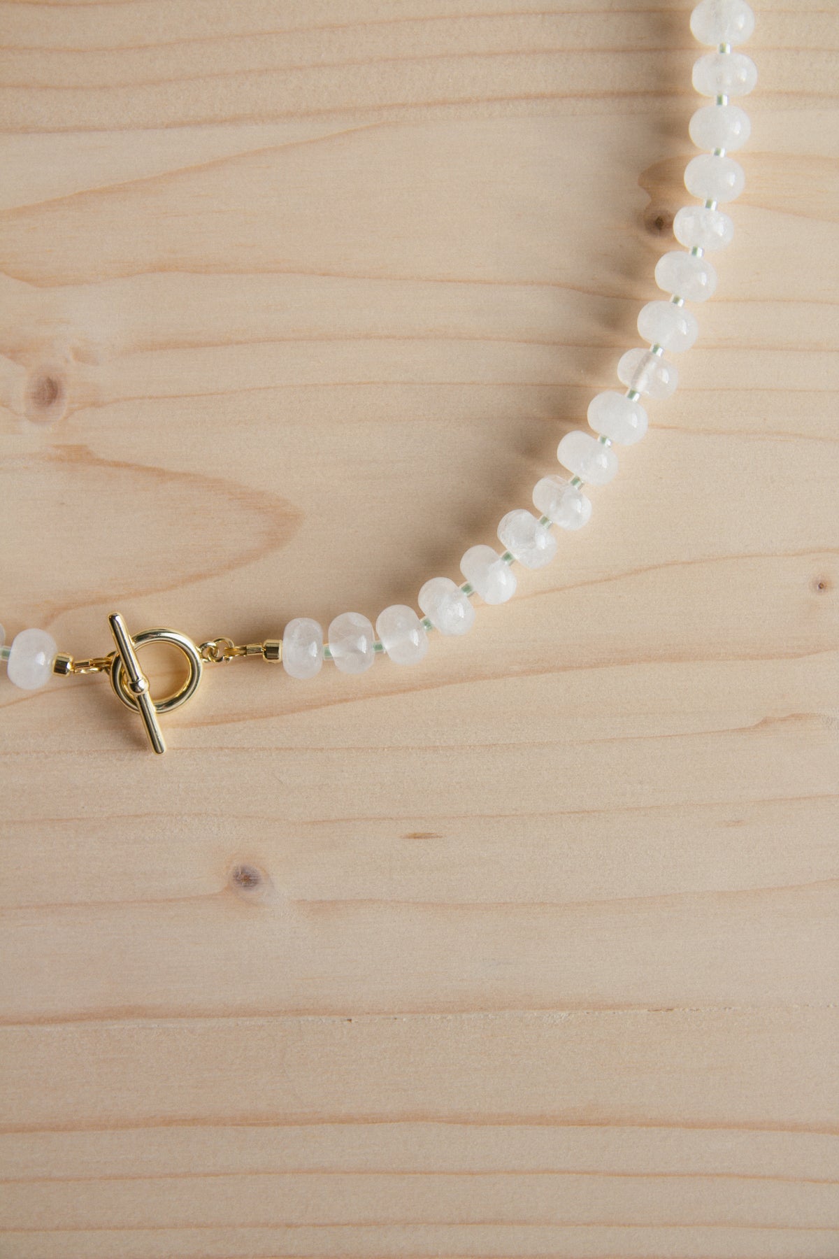 A beaded necklace made of small white oval beads with a gold toggle clasp, laid on a light wood surface.