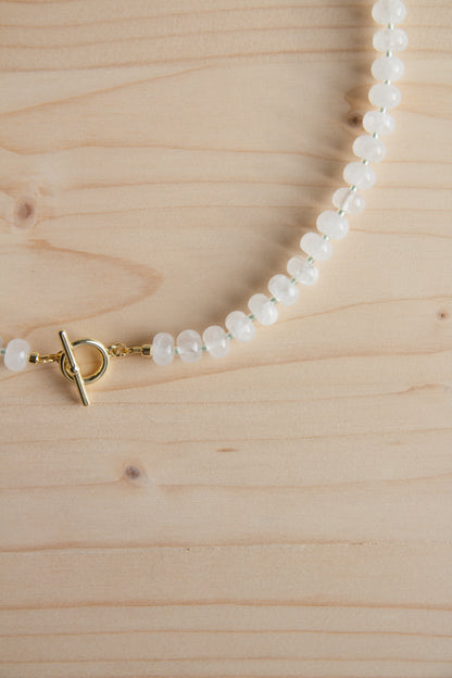 A beaded necklace made of small white oval beads with a gold toggle clasp, laid on a light wood surface.