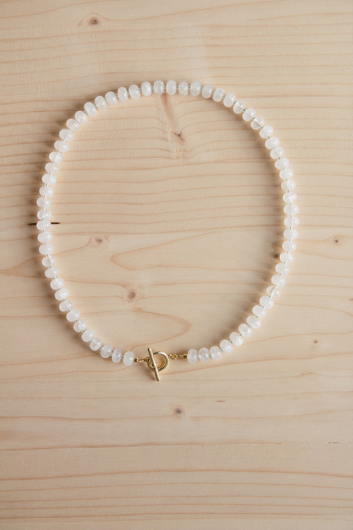 A beaded necklace made of small white oval beads with a gold toggle clasp, arranged in a circle on a light wood grain background.