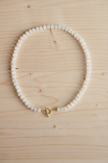 A beaded necklace made of small white oval beads with a gold toggle clasp, arranged in a circle on a light wood grain background.