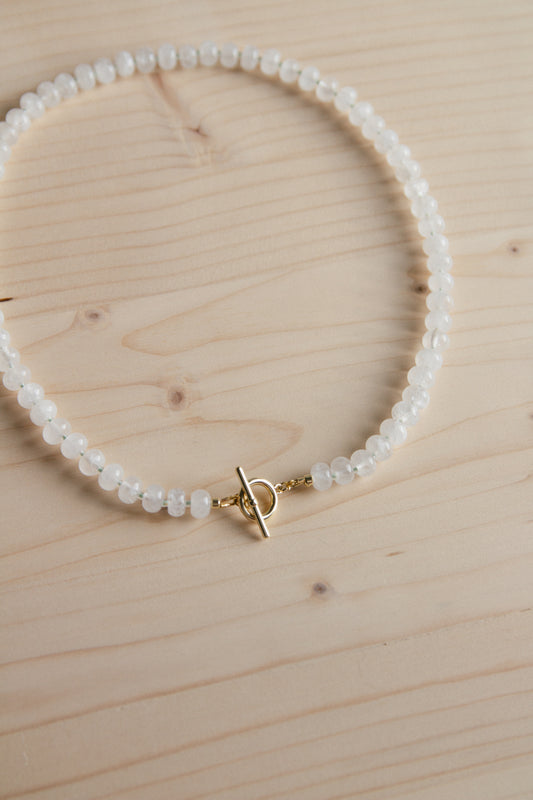 A beaded necklace made of small white faceted beads with a gold toggle clasp, displayed on a light wood grain surface.